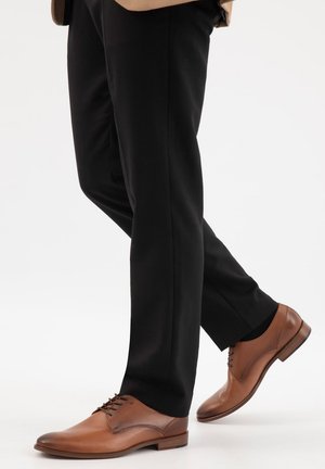 Mann trägt braune Lederschuhe und schwarze Hose, geht mit einem Fuß nach vorne auf weißem Hintergrund.