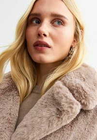 Cappotto in pelliccia sintetica di un beige chiaro, con un colletto spesso e una texture morbida. Il modello indossa un top beige e orecchini ad anello dorati.