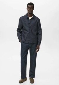 Donkerblauwe denim knoopjesjas gecombineerd met bijpassende broeken. De stof heeft een gladde textuur. De outfit wordt aangevuld met bruine schoenen.