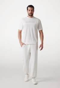 White cotton t-shirt with "GUESS" logo on front, paired with matching white joggers featuring pockets and cuffed hems. White sneakers complete the look.