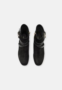 Paire de bottines en daim noir à bouts carrés, avec des lanières en cuir décoratives enveloppantes et des boucles dorées, vues de dessus.