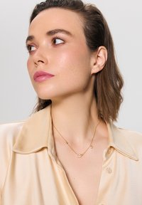 Złoty naszyjnik z sercowym wisiorkiem; delikatny łańcuszek z małym akcentem w postaci perełki; modelka nosi jasną satynową koszulę.