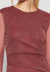 Burgundy topp med strukturerade metalltrådar, långa genomskinliga ärmar och en design med böjda sömmar. Slät textil med subtil lyster.