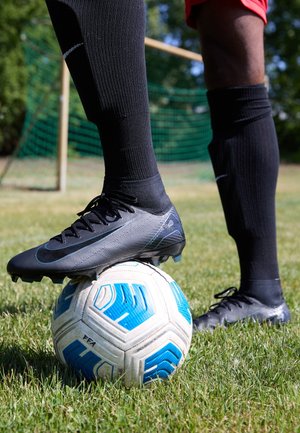 Des crampons de football noirs reposent sur un ballon de football blanc et bleu. Terrain de gazon en arrière-plan, un joueur porte des chaussettes noires et un short rouge.