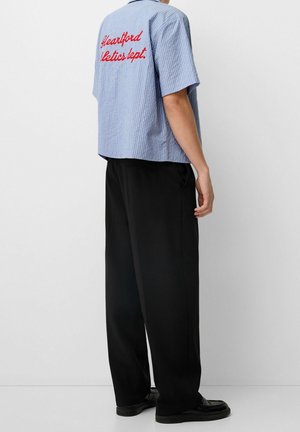 Mężczyzna w niebieskiej koszuli w paski z krótkim rękawem z czerwonym napisem „Heartford Athletics dept.” na plecach, czarne spodnie z szerokimi nogawkami i czarne buty.