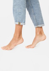 Náhrdelník na holé noze; džínsy stočené na manžetách. Svetlomodrý denim, voľný strih, odhaľujúci členok so zlatým reťazovým detailom a jemnou textúrou pleti.