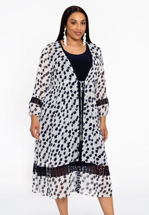Robe blanche transparente avec des taches bleu foncé et noires, manches trois-quarts, taille nouée et détail en treillis contrastant au niveau de l'ourlet.