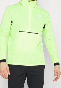 Chaqueta ligera de color verde neón con media cremallera, detalles en negro y puños elásticos. Bolsillo con cremallera en el frente y capucha integrada para mayor funcionalidad.