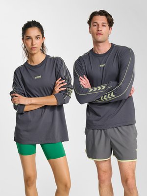 Zwei Erwachsene stehen mit verschränkten Armen, tragen passende dunkelgraue Langarmshirts mit hellgrünen Akzenten und Shorts, neutraler Hintergrund.