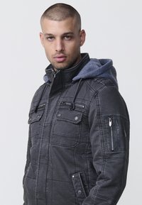 Mörkgrå jacka med quiltat m�önster, en blå hoodie, flera blixtlås- och knappfickor, samt synliga söm detaljer.
