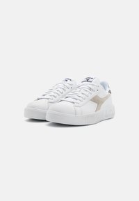 Baskets blanches en cuir avec un col rembourré, présentant une touche beige sur le côté, une semelle en caoutchouc texturée et des lacets plats.