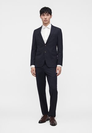 Jeune homme portant un costume bleu marine foncé avec une chemise blanche et des chaussures marron, debout devant un fond clair uni.