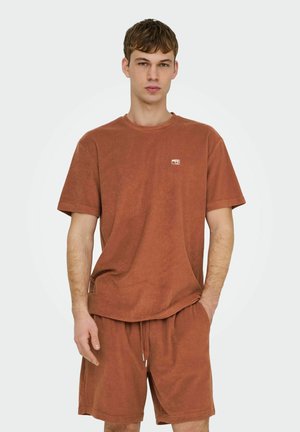 Jonge man die een roestkleurig T-shirt met korte mouwen en bijpassende korte broek draagt, met één hand in de zak, staand tegen een effen achtergrond.