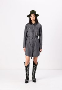 Robe chemise en denim gris avec manches longues, devant boutonné, deux poches poitrine et ceinture à la taille ; assortie avec des bottes de cowboy noires embellies.