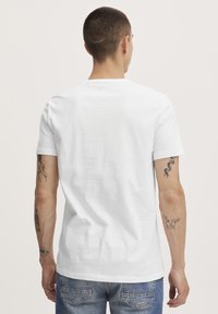 Valge puuvillane T-särk ümarlõikega kaeluse, lühikeste varrukate ja ühtlase tekstuuriga, nähtav tagant poolt, sobitatud siniste teksapükstega.