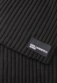 Tecido de malha ribada preto com uma textura tecida, apresentando uma etiqueta retangular com o texto "KARL LAGERFELD JEANS" em branco sobre um fundo preto.