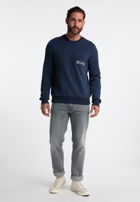 Felpa blu navy in misto cotone, con tasca frontale e logo ricamato, abbinata a jeans grigi chiari e sneakers bianche.