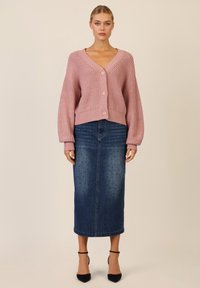 Różowy sweter dzianinowy z dekoltem w kształcie litery V i guziczkami, zestawiony z dżinsową spódnicą midi w kropki. Całość uzupełniają szpilki.