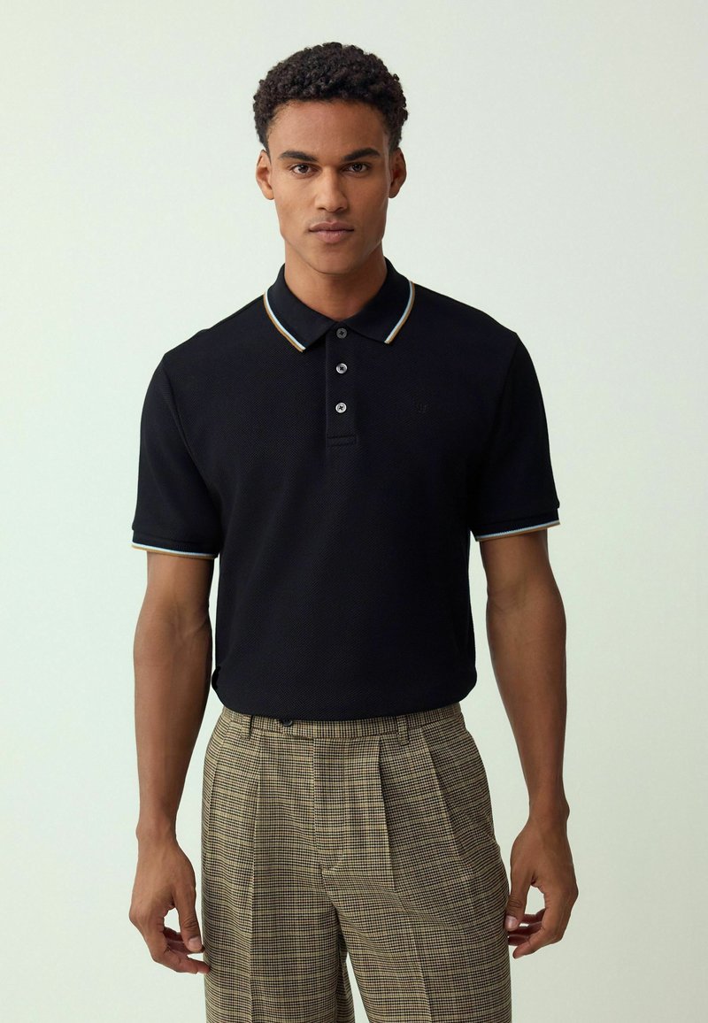 Schwarzes Poloshirt aus strukturiertem Stoff, mit einem Kragen mit mehrfarbigen Streifen und drei Knöpfen. Kombiniert mit gemusterten braunen Hosen.