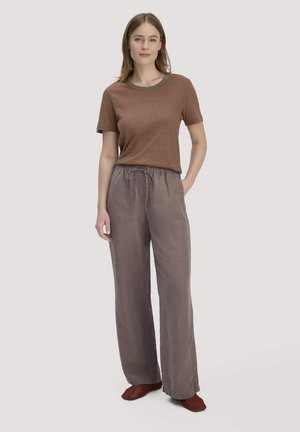 Frau steht mit einer Hand in der Hosentasche, trägt ein braun gestreiftes T-Shirt, lockere graue Hosen und rote gewebte Slipper.