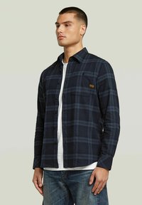 Camicia di flanella a quadri blu navy con chiusura a bottoni, maniche lunghe, due tasche sul petto e un piccolo marchio sulla tasca sinistra.