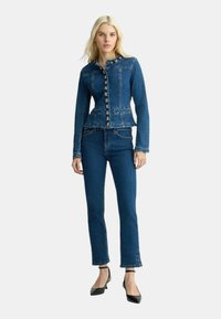 Giacca in denim con un design aderente, colletto rotondo e chiusura con bottoni. Colore blu scuro con cuciture chiare e due tasche frontali, abbinata a jeans a gamba dritta.