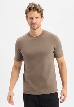 Man with curly hair wearing a plain brown short-sleeve t-shirt and black pants, standing against a white background.