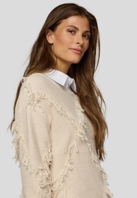 Beiger Pullover mit strukturierten Fransen entlang der Nähten, mit Rundhalsausschnitt. Weißer Hemdkragen ist darunter sichtbar.