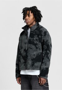 Giacca in pile nei colori nero e grigio con un motivo testurizzato, collare a mezza zip e branding discreto sul petto.