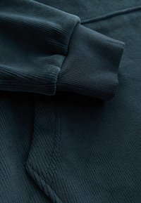 Tessuto di un verde scuro con texture a righe e polsini lisci. Primo piano di una felpa che mostra una cucitura dettagliata e una vestibilità rilassata.