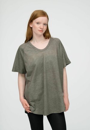 Giovane donna con capelli rossi lisci indossa una camicia larga verde oliva a maniche corte e leggings neri, in piedi davanti a uno sfondo chiaro e uniforme.