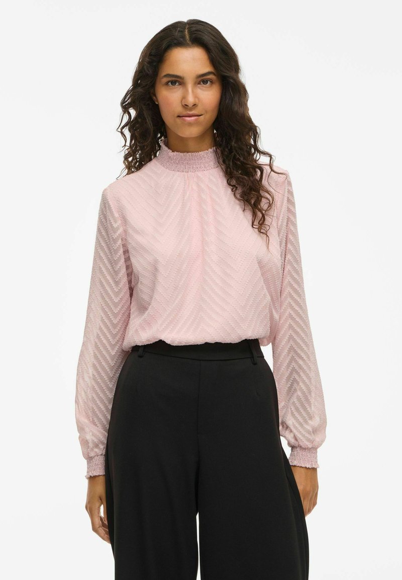 Rosa långärmad blus med zigzag-mönster, hög smockad halsringning och texturerad tyg, tillsammans med svarta högmidjade byxor.