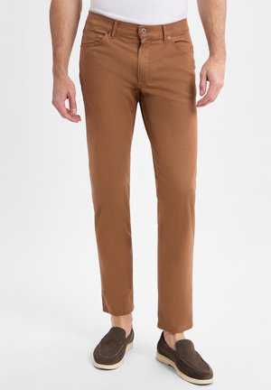 Braune Baumwollhose im Slim-Fit-Stil, mit Reißverschluss, Gürtelschlaufen und vorderen Taschen, kombiniert mit dunkelbraunen Slippern.