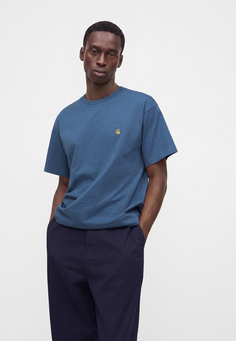 Junger Mann mit dunkler Haut, der ein blaues T-Shirt und dunkle Hose trägt, steht mit den Händen in den Taschen vor einem schlichten weißen Hintergrund.