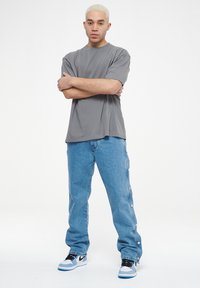 Camiseta gris de manga corta combinada con unos jeans de mezclilla azul claro que tienen botones a presión a lo largo de los lados y zapatillas deportivas en blanco y negro.