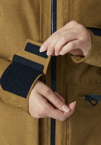 Chaqueta texturizada marrón con acentos de velcro negro en los puños. Dos dedos ajustando el cierre de velcro. Bolsillo con cremallera visible en el lado.