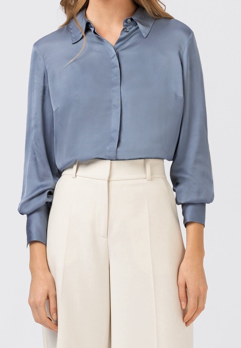 Kvinna iklädd en glänsande blå långärmad blus som är instoppad i högmidjade, crèmefärgade byxor med vida ben framför en enfärgad bakgrund.