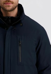Mann in einer marineblauen Jacke mit einer Reißverschlusstasche auf der Brust und hohem Kragen über einer schwarzen Reißverschlussjacke vor einem einfarbigen Hintergrund.