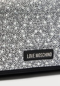 Μαύρο τσαντάκι χειρός διακοσμημένο με διάφορα μεγέθη διαφανών στρας· διαθέτει υφή στην επιφάνεια και μαύρη δερμάτινη πλάκα με το λογότυπο.