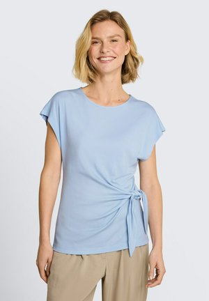 Vrouw glimlacht, draagt een lichtblauw shirt met korte mouwen en een knoop aan de zijkant, gecombineerd met beige broek tegen een effen achtergrond.