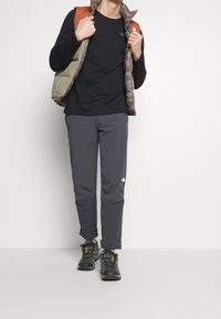 Herren Outdoor-Outfit mit einem schwarzen Langarmshirt, einer olivgrünen gepolsterten Weste mit Camouflage, dunkelgrauen Hosen und grünen Wanderschuhen.