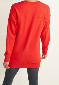 Personne portant un pull à manches longues rouge vif avec des poignets et un ourlet côtelés, associé à un jean en denim foncé, vue de dos.