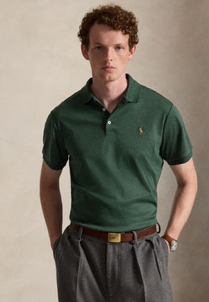 Jeune homme aux cheveux roux bouclés portant un polo vert foncé et un pantalon gris avec une ceinture marron, debout les mains dans les poches.