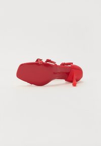 Chaussure à talon rouge avec une finition lisse et brillante. Présente un talon mince et des détails de sangle autour de la cheville, avec une semelle texturée pour l'adhérence.