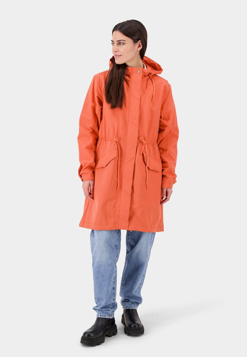 Vrouw die staat in een oranje, met een capuchon uitgeruste regenjas met trekkoorden, lichte spijkerbroek en zwarte chunky laarzen op een effen achtergrond.