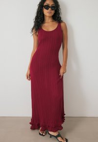 Rode geribde maxi-jurk met een ronde hals en een rucheszoom. Draag met zwarte zonnebril en instapsandalen. Glad textuur, aansluitend ontwerp.