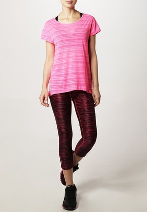 Roze sportshirt met verticale strepen, gecombineerd met zwarte en rode capri-legging met patroon. Model draagt zwarte sneakers.