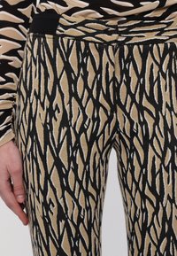 Gros plan sur un pantalon beige et noir à motif abstrait porté avec un haut à manches longues assorti, montrant la main gauche détendue sur le côté.