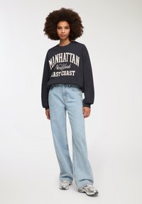 Felpa blu navy con stampa bianca "MANHATTAN New York EAST COAST," abbinata a jeans a gamba larga blu chiaro e sneakers argentati.