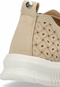 Zapatilla de ante beige con patrones de corte circular, suela de goma blanca y un tirador cosido. Presenta un acento metálico cerca de la parte superior.